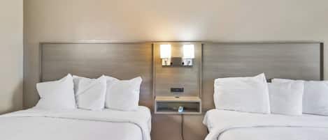 A hotel room with two beds, each with white bedding and pillows. There is a wall-mounted light fixture between the beds.