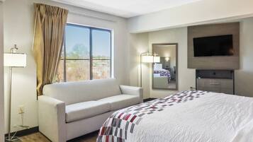 A hotel room with a bed, a sofa, a TV, a window with curtains, and a bedside lamp.