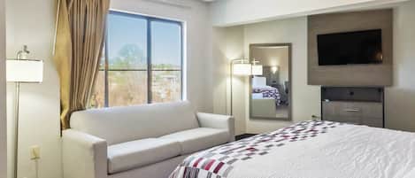 A hotel room with a bed, a sofa, a TV, a window with curtains, and a bedside lamp.