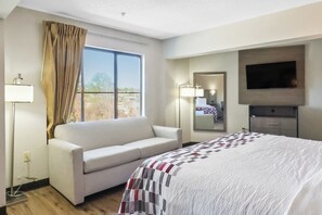 A hotel room with a bed, a sofa, a TV, a window with curtains, and a bedside lamp. - Red Roof Inn & Suites Fayetteville - Fort Bragg (Fayetteville)
