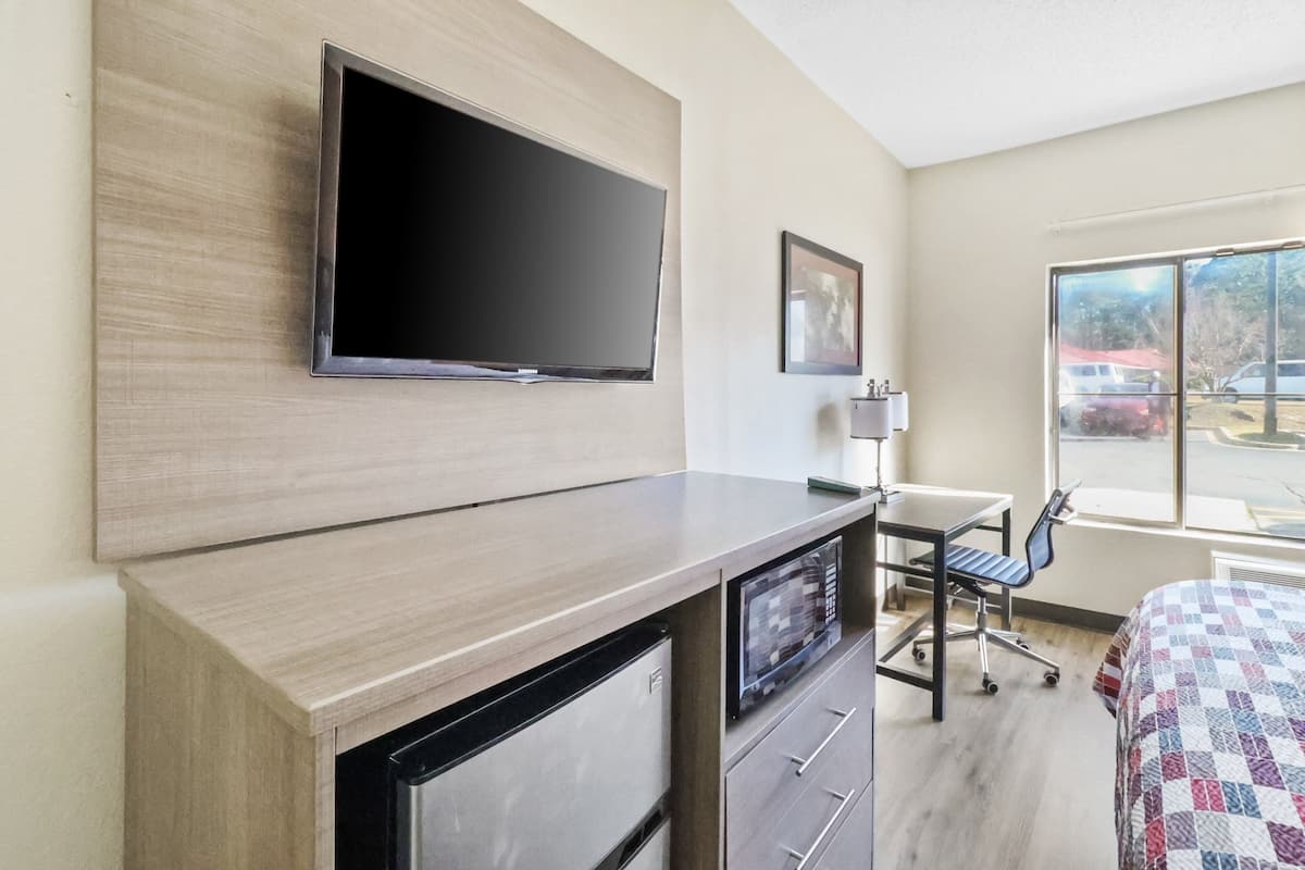 A hotel room with a flat-screen TV mounted on the wall, a desk with a chair, a bed with a checkered bedspread, and a window with a view of parked cars outside.