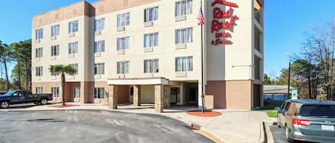 A multi-story hotel with a beige facade, a red sign reading "Red Roof Inn," and a parking lot with several vehicles.