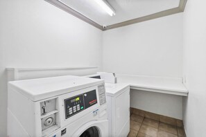A laundry room with a front-loading washing machine and dryer, a control panel, and a shelf above the dryer. - Red Roof Inn & Suites Fayetteville - Fort Bragg (Fayetteville)