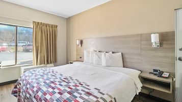 A hotel room with a bed, a nightstand, a window with curtains, and a view of parked cars outside.