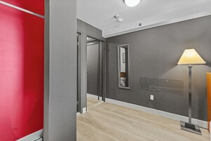 A hallway with a red wall, a grey wall, and a wooden floor. There is a mirror on the grey wall and a lamp on the right side.
