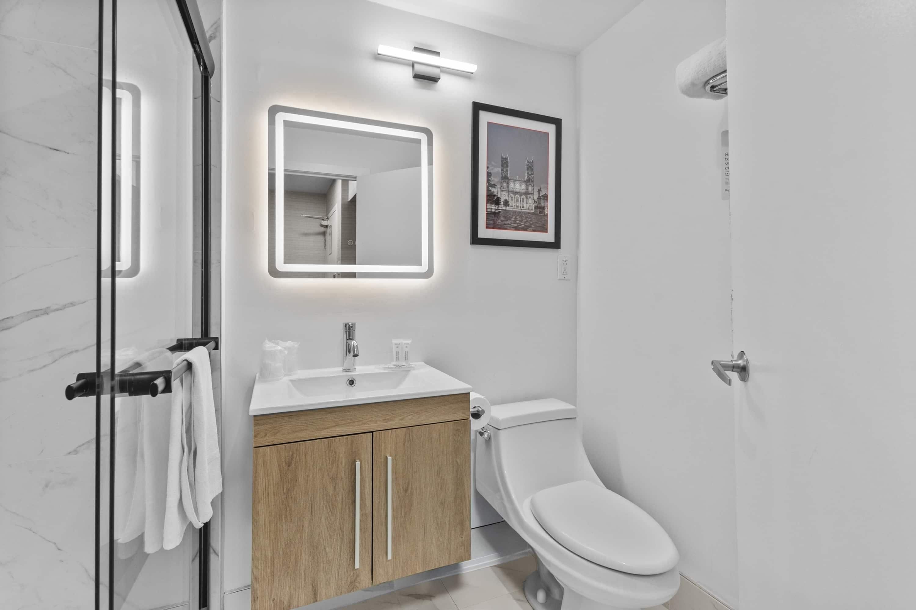 Un baño moderno con cabina de ducha de vidrio, tocador de madera, inodoro blanco y un cuadro en la pared.