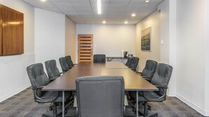 Un salón de conferencias con una larga mesa, sillas negras, paredes blancas, una puerta de madera y un cuadro en la pared.