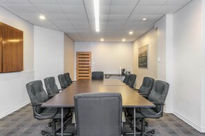 Un salón de conferencias con una larga mesa, sillas negras, paredes blancas, una puerta de madera y un cuadro en la pared.