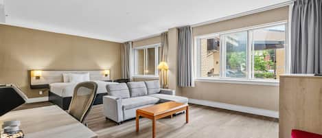 Une chambre d’hôtel comprenant un lit, un canapé, une table en bois, un bureau avec une chaise, une lampe et de grandes fenêtres avec des rideaux.
