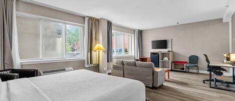 A hotel room with a bed, a desk, a chair, a TV, and large windows.