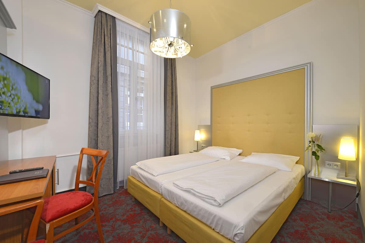 Una habitación de hotel con cama, escritorio con silla, televisión y mesita de noche con lámpara.