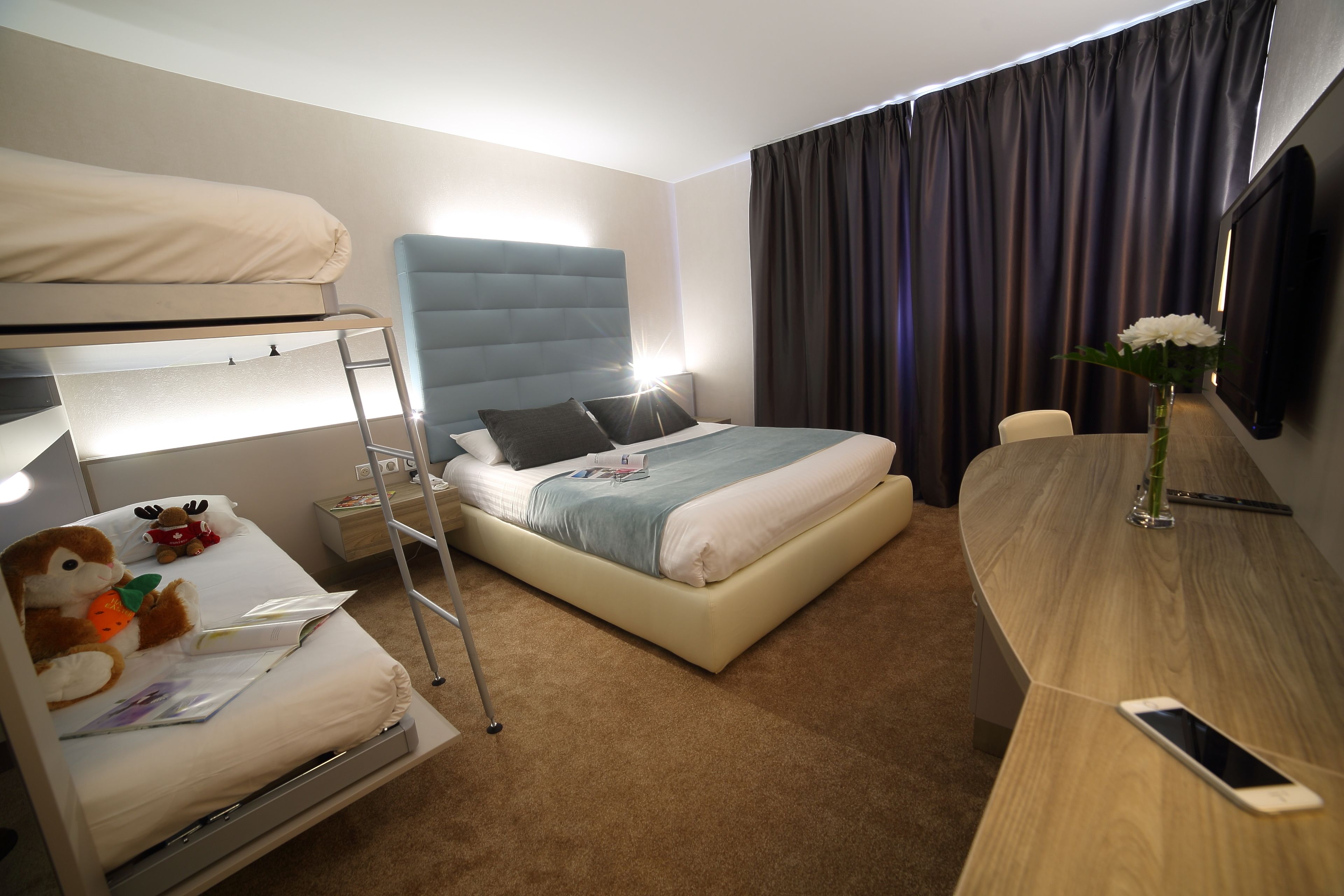 A hotel room with a bunk bed, a desk, a chair, a lamp, a mirror, and a vase with flowers.