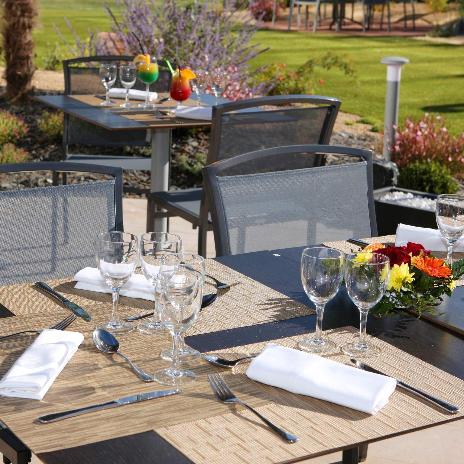 An outdoor dining area with tables set for two, complete with cutlery, glasses, and a flower arrangement.