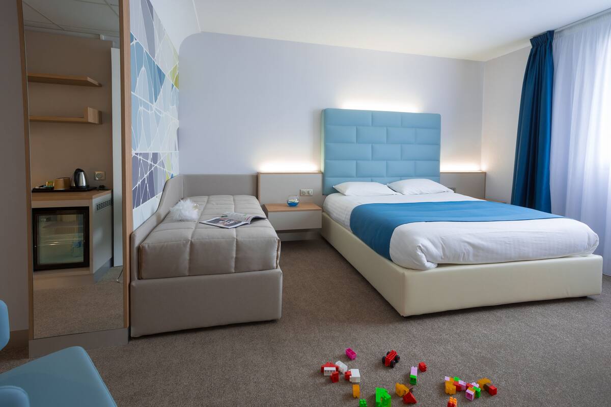 Ein Hotelzimmer mit einem großen Bett, einem kleineren Sofa, einem Schreibtisch mit Wasserkocher und Spielzeug auf dem Boden.