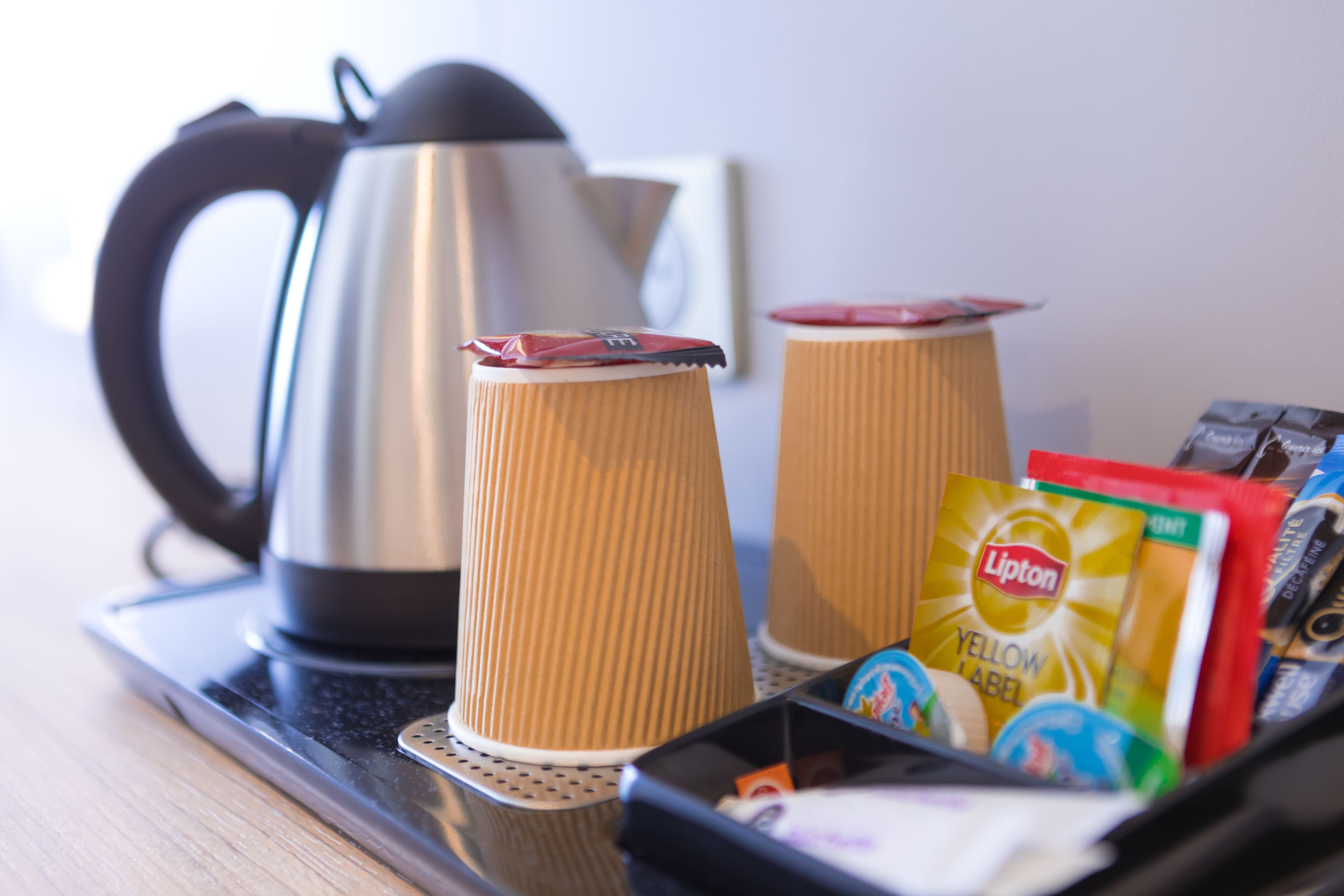 Una bandeja con un hervidor de acero inoxidable, dos vasos de papel, un sobre de té Lipton y una pequeña caja de pañuelos.