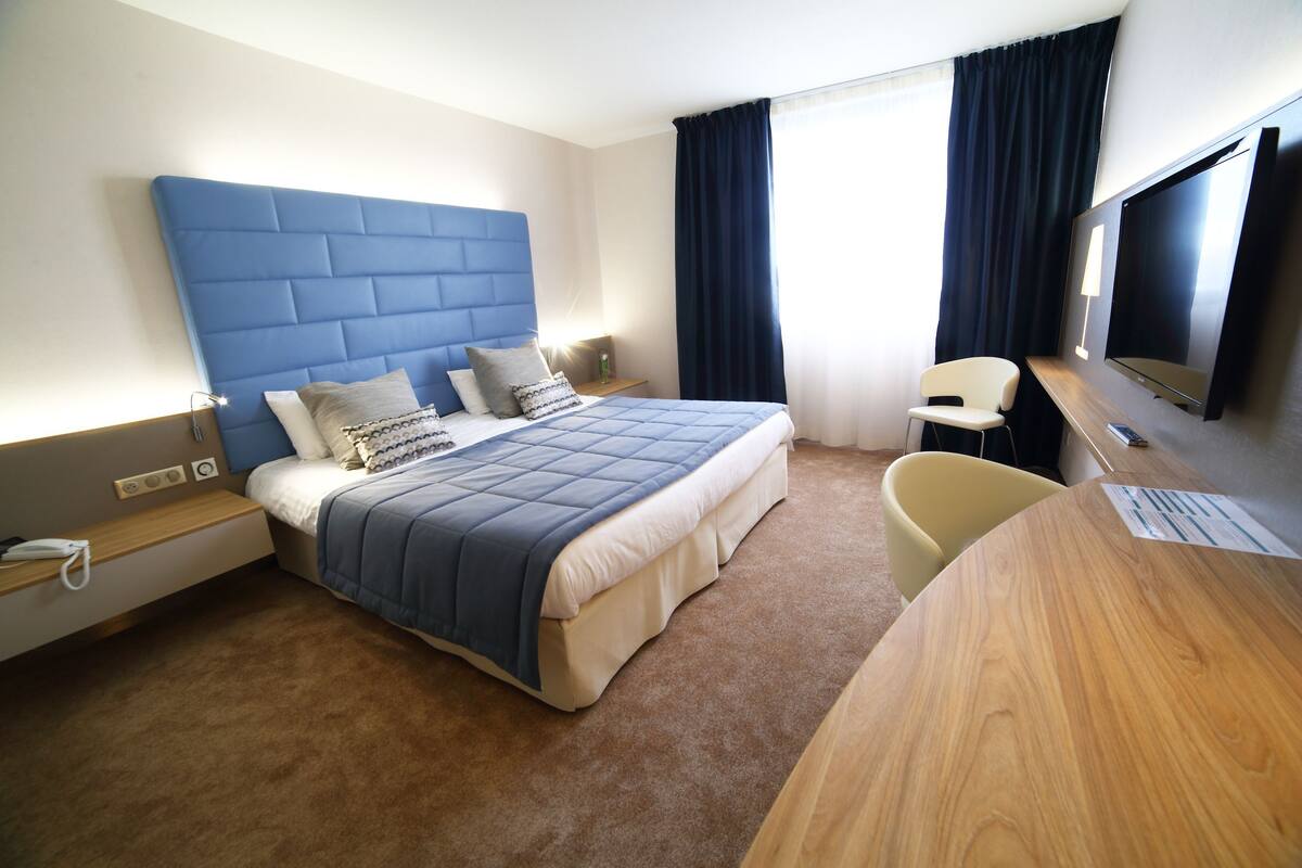 Ein Hotelzimmer mit einem großen Bett, einem Schreibtisch, einem Stuhl, einem Fernseher und einem Vorhangfenster.