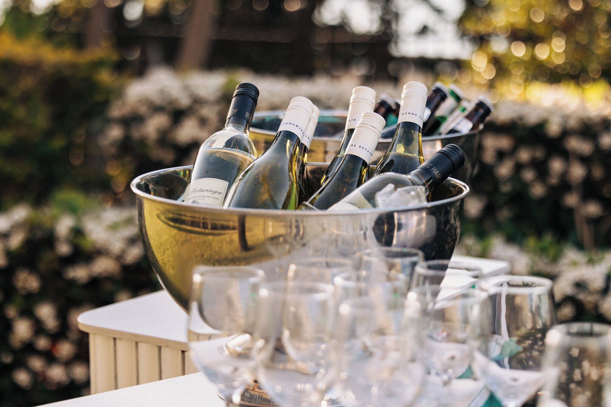 Un seau en métal rempli de bouteilles de vin et de verres, posé sur une table à l’extérieur.