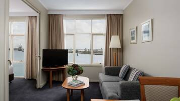 A hotel room with a large window, a flat-screen TV, a sofa, a coffee table, and a dining table with apples.
