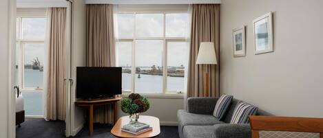 A hotel room with a large window, a flat-screen TV, a sofa, a coffee table, and a dining table with apples.