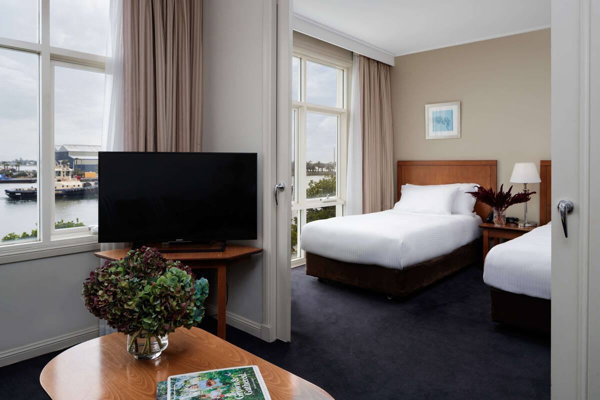 Hotellihuone, jossa on sänky, televisio, ikkuna, josta on näkymä vesistöön, sekä pöytä, jonka päällä on maljakko täynnä kukkia.