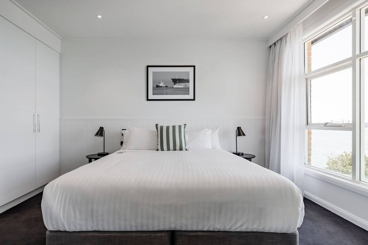 Moderni hotellihuone, jossa on suuri sänky, valkoiset seinät, seinällä oleva taulu ja ikkunasta avautuva merinäköala.