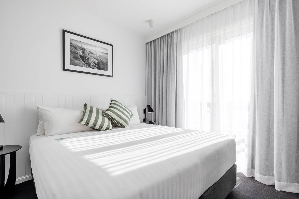 Hotellihuone, jossa on suuri sänky, yöpöytä, lamppu, seinällä oleva kehystetty kuva ja läpikuultavat verhot ikkunassa.