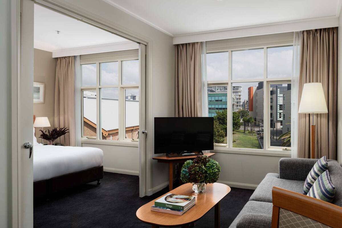 Hotellihuone, jossa on sänky, televisio, sohva, kirjoja sisältävä sohvapöytä ja suuret ikkunat, joista avautuu näkymä kaupunkiin.