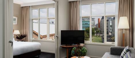 A hotel room with a bed, a TV, a sofa, a coffee table with books, and a view of the city through large windows.