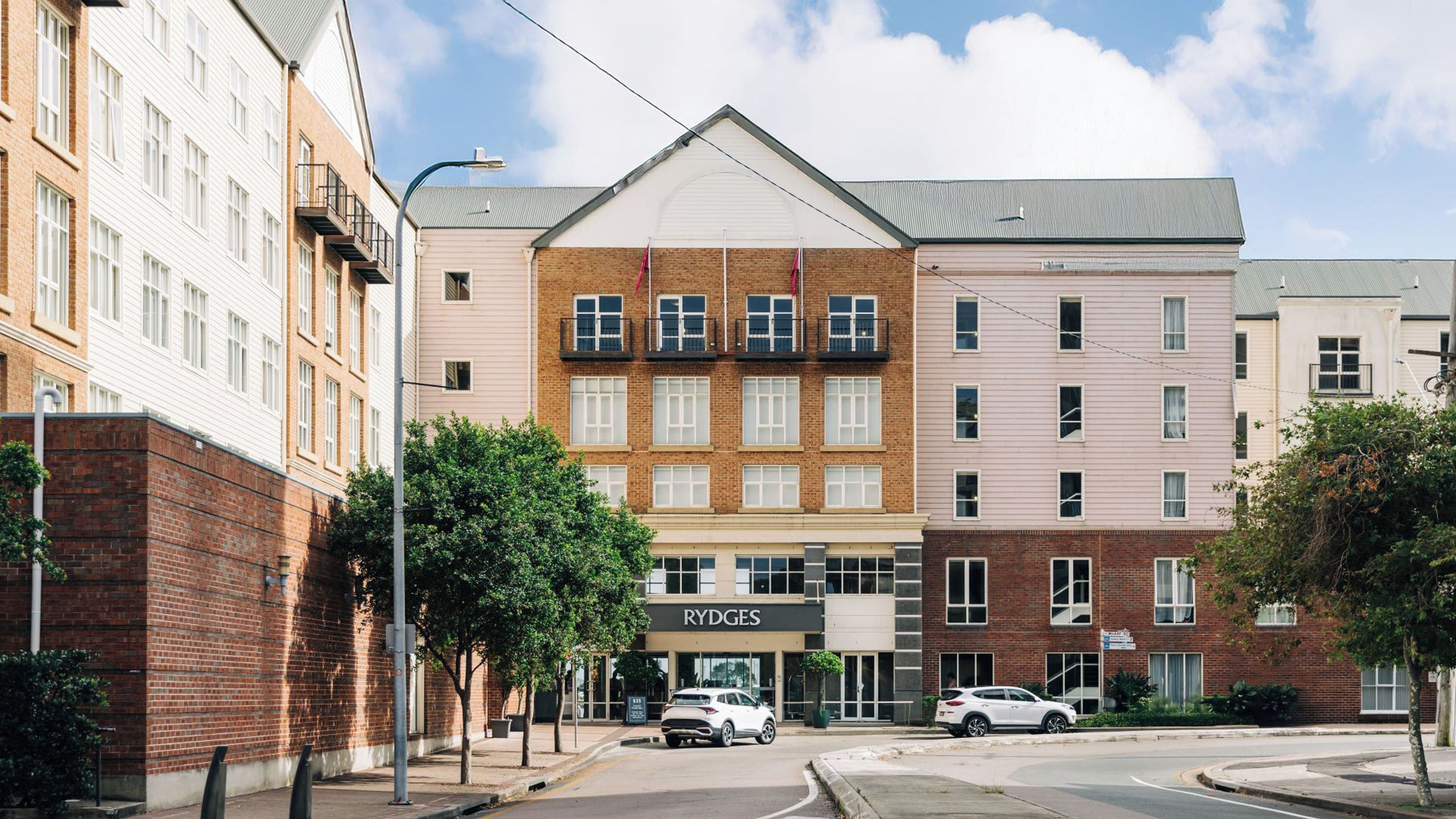 A multi-story building with a central entrance, a sign reading 'RYDGES,' and two cars parked in front.