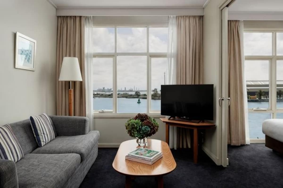 A hotel room with a sofa, a coffee table with a book and a plant, a TV on a stand, and a view of a body of water through large windows.