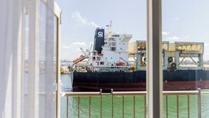 Un vaixell de càrrega gran amarrat en un port, amb grues i construccions industrials al fons.