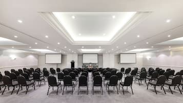 A conference room with rows of chairs facing a podium, a large screen, and a ceiling with recessed lighting.