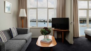 Habitació d'hotel amb un sofà, una taula de café amb un llibre i un vas, una televisió i grans finestres amb vista a un cos d'aigua i un pont.