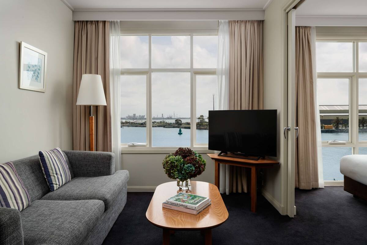 Hotellihuoneessa on sohva, sohvapöytä, jonka päällä on kirja ja maljakko, televisio sekä suuret ikkunat, joista avautuu näkymä vesistöön ja sillalle.