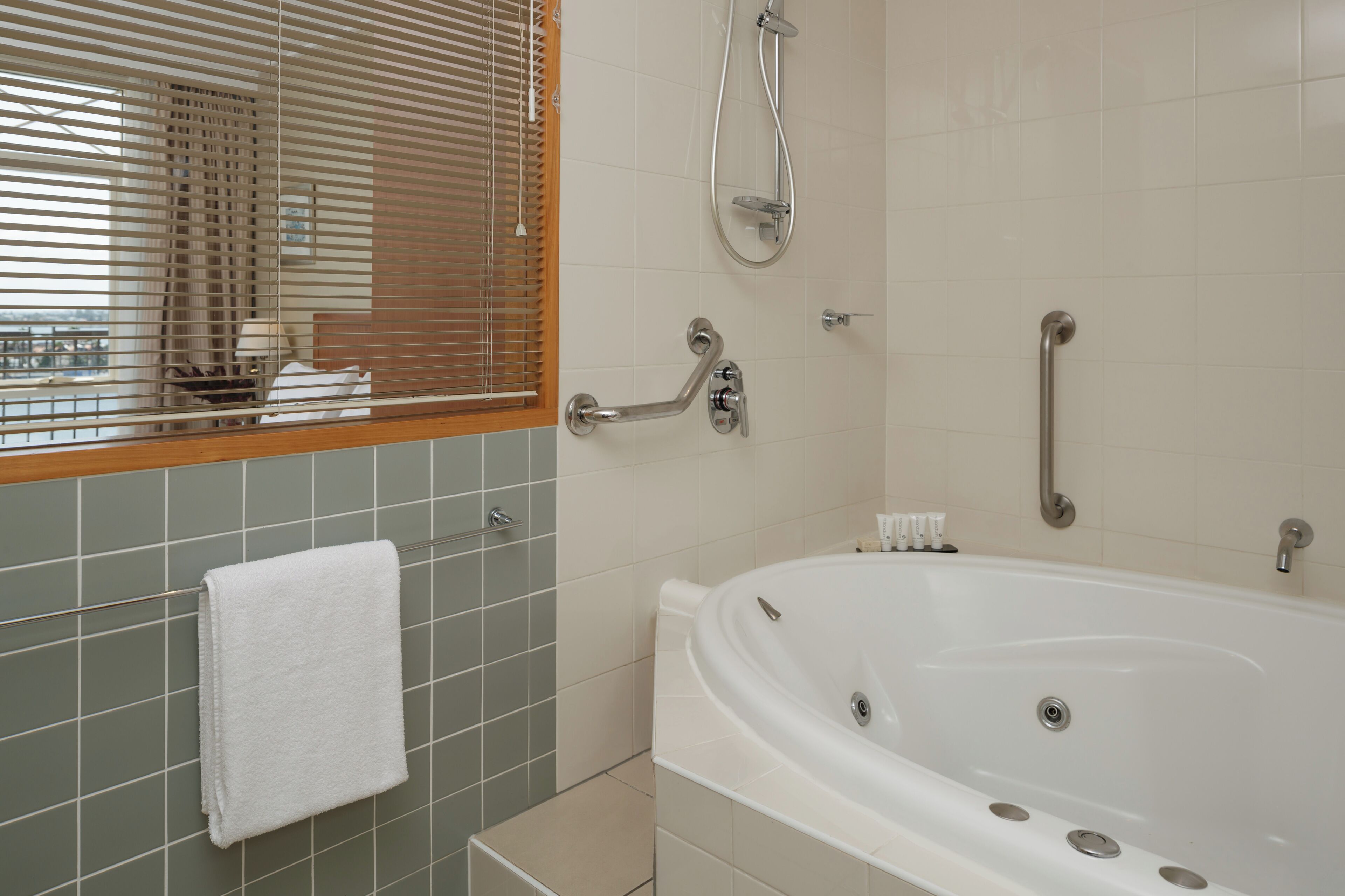 Un baño con bañera, ducha, barra de apoyo, toallero y ventana con persianas.