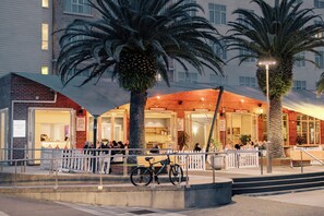 A hotel with a covered outdoor seating area, palm trees, and a bicycle parked outside.