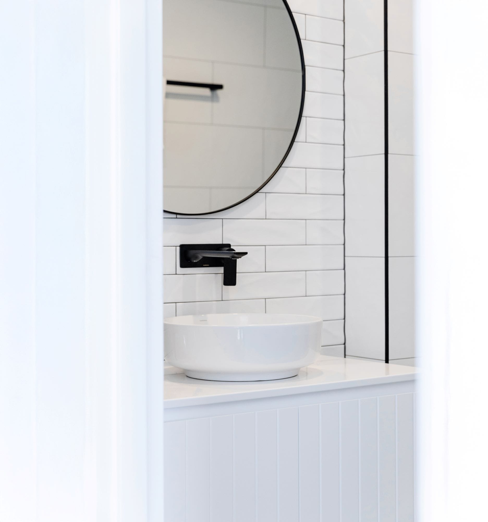 A white bathroom with a round mirror, a black faucet, and a white vessel sink.
