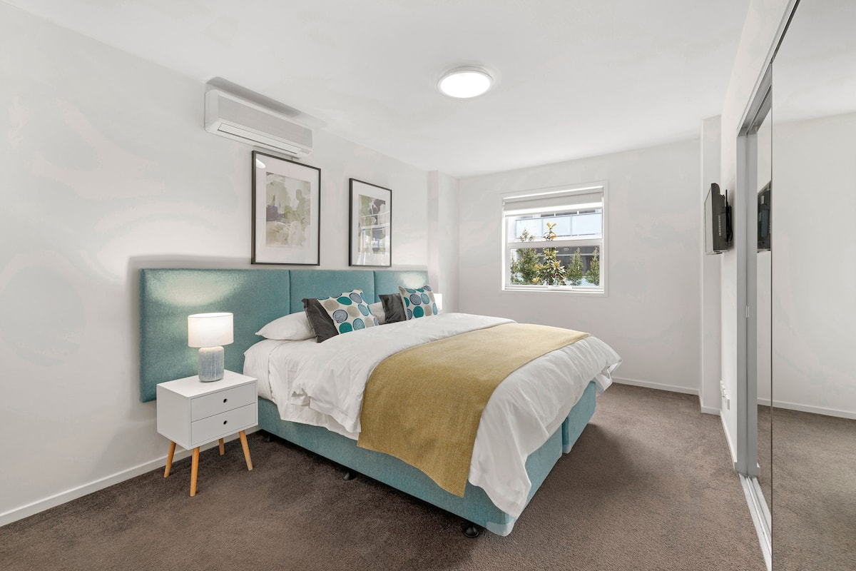 Une chambre avec un lit de couleur turquoise, du linge de lit blanc, une lampe de chevet et une fenêtre donnant sur un paysage verdoyant.