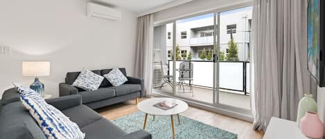Uma sala de estar moderna com um sofá cinzento, uma mesa de centro redonda e vista para uma varanda com mobiliário de exterior.