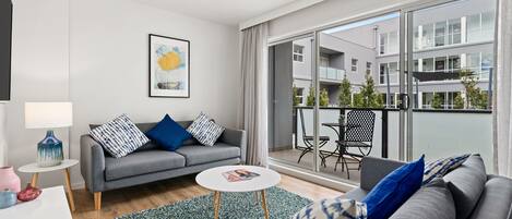 Ein modernes Wohnzimmer mit einem grauen Sofa, einem kleinen runden Tisch und einem Balkon mit Blick auf andere Gebäude.