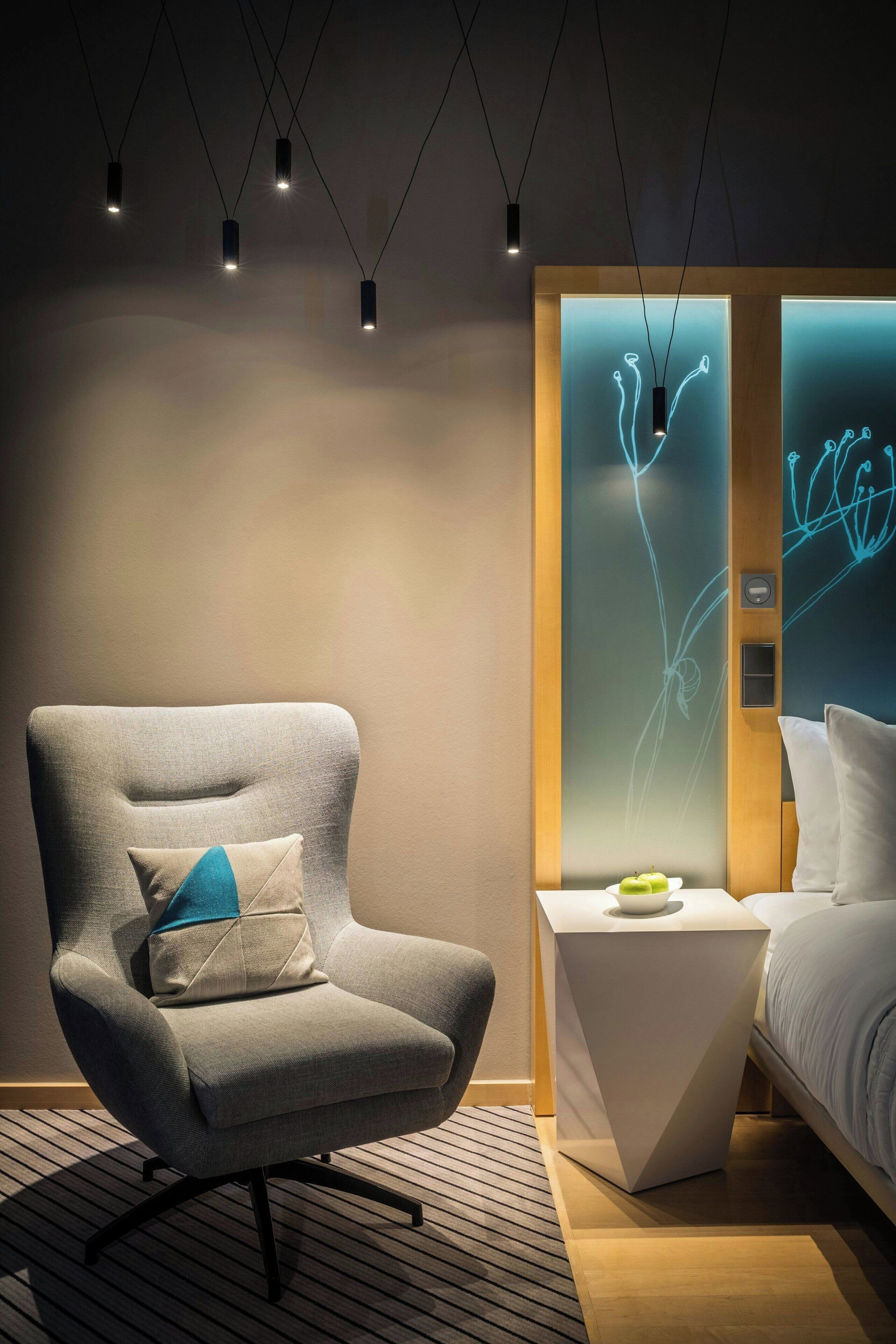 Una habitación de hotel moderna con un sillón gris, una mesita de noche con un objeto verde y una cama con ropa blanca.