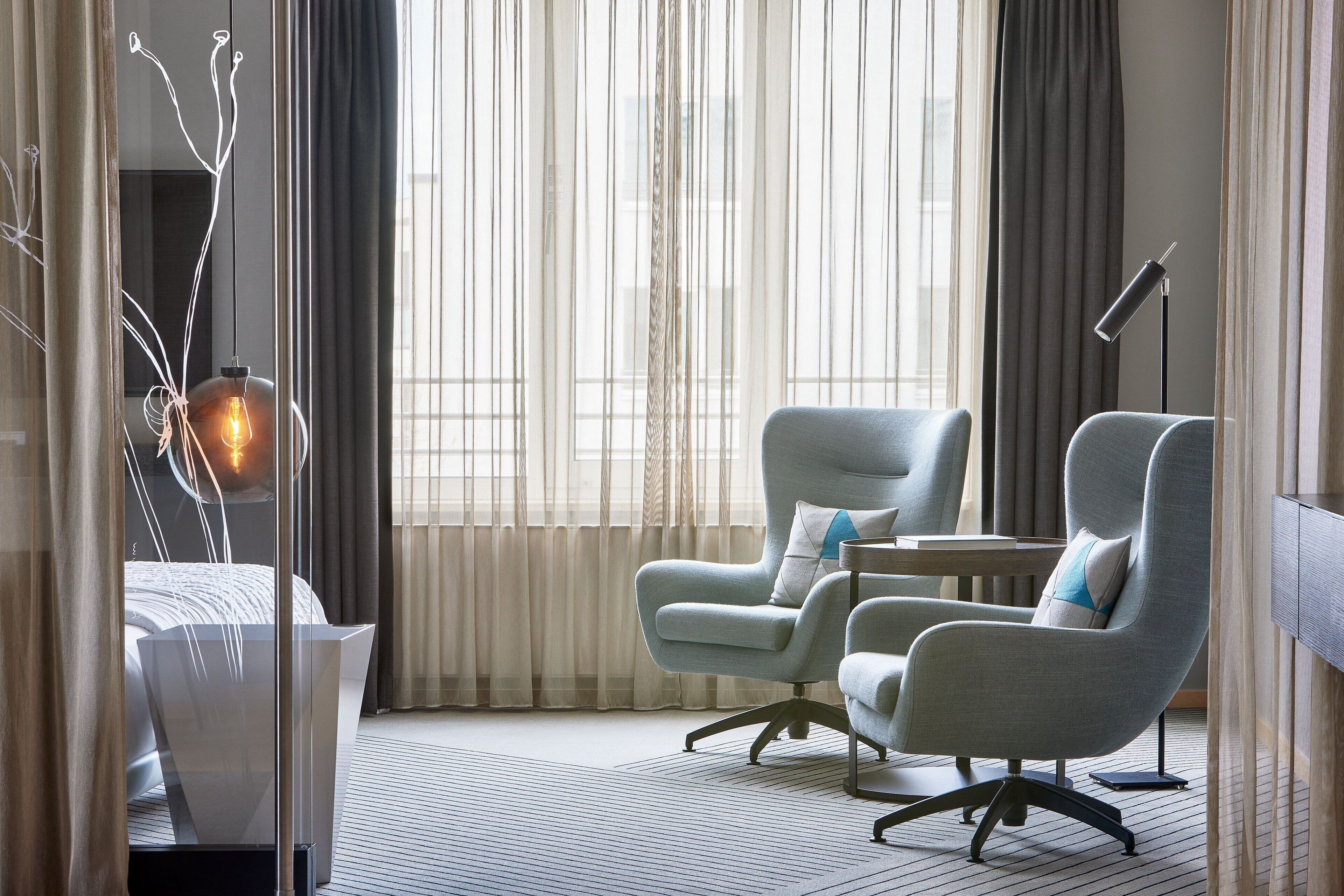 Una habitación de hotel moderna con un gran ventanal, dos sillones, un escritorio y una lámpara de pie.