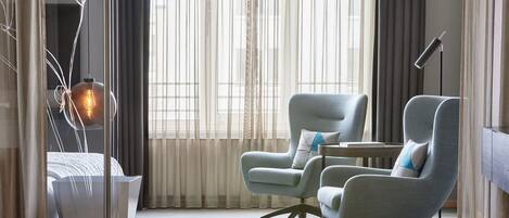 Una habitación de hotel moderna con un gran ventanal, dos sillones, un escritorio y una lámpara de pie.