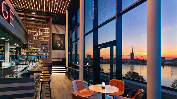 A modern bar with a long counter, stools, and a large window overlooking a cityscape at sunset. - Le Méridien Hamburg (Hamburg)