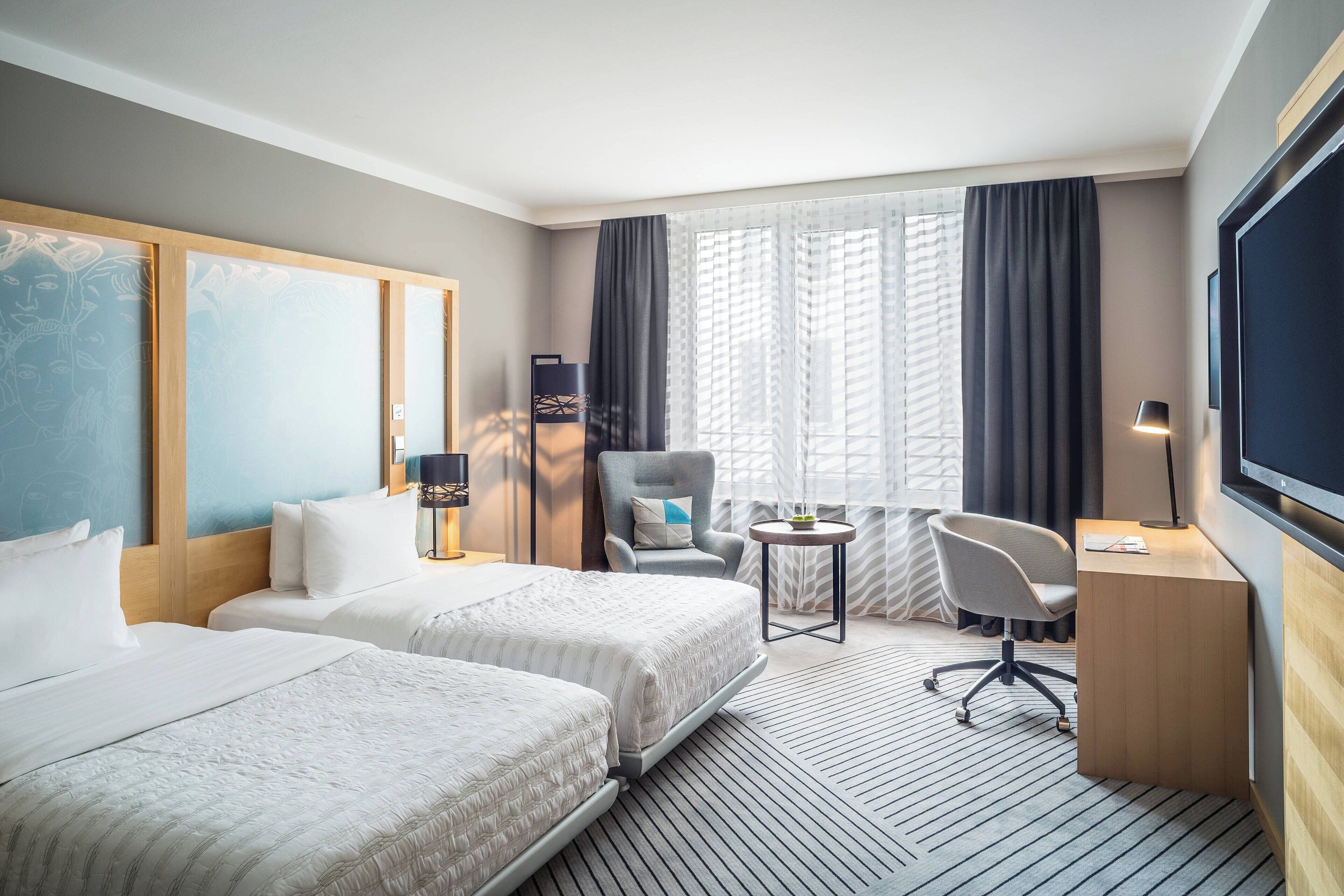 Ein Hotelzimmer mit zwei Betten, einem Schreibtisch, einem Stuhl, einem Fernseher und großen Fenstern mit Vorhängen.