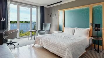 Ein Hotelzimmer mit einem großen Bett, einem Schreibtisch, einem Sessel, einem kleinen Tisch und Blick auf das Wasser.