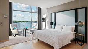A modern hotel room with a large bed, a seating area with a view of the water, and a balcony with a railing. - Le Méridien Hamburg (Hamburg)