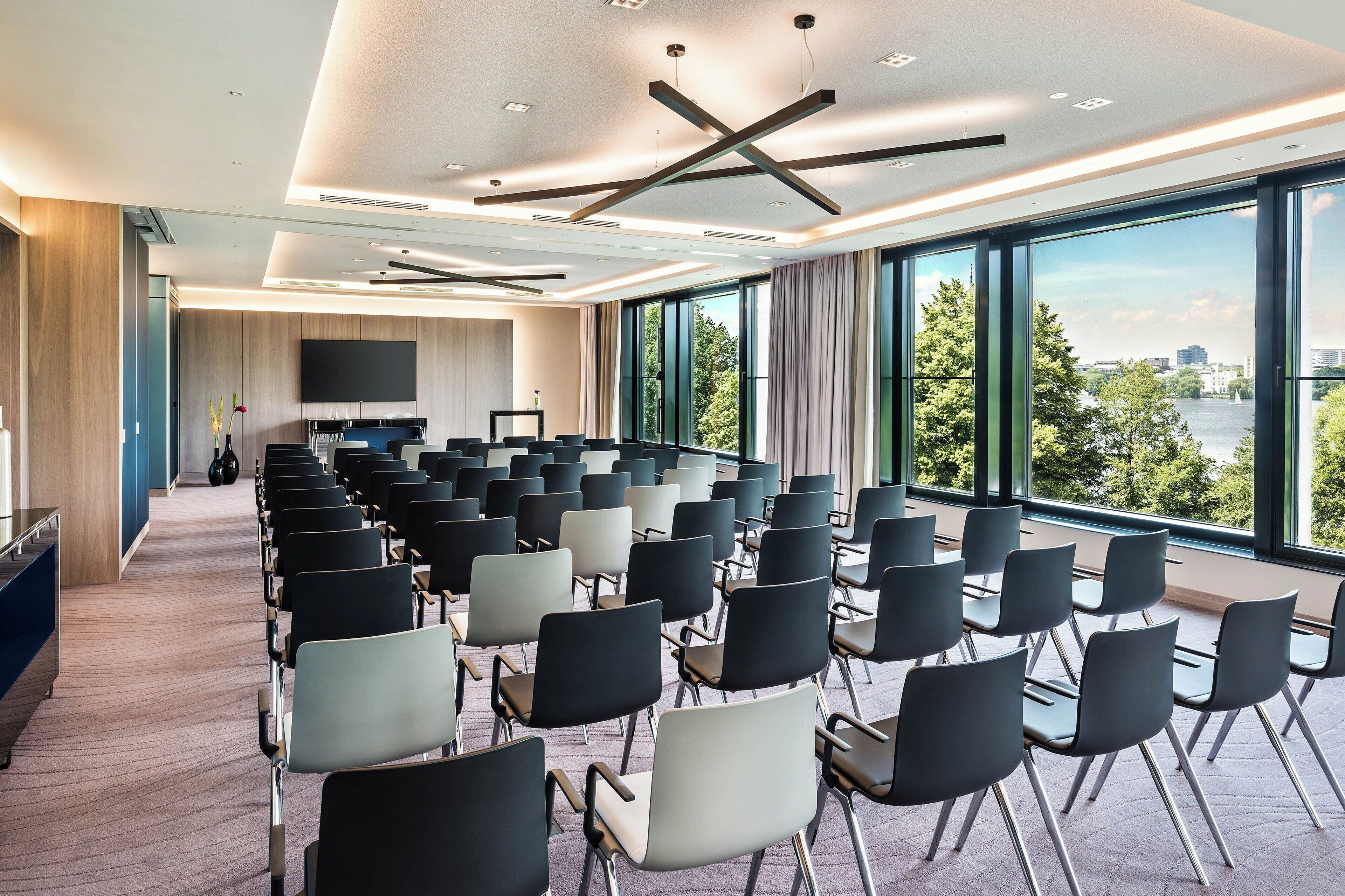 A conference room with rows of chairs, a large TV screen, and a view of a river and cityscape through large windows.