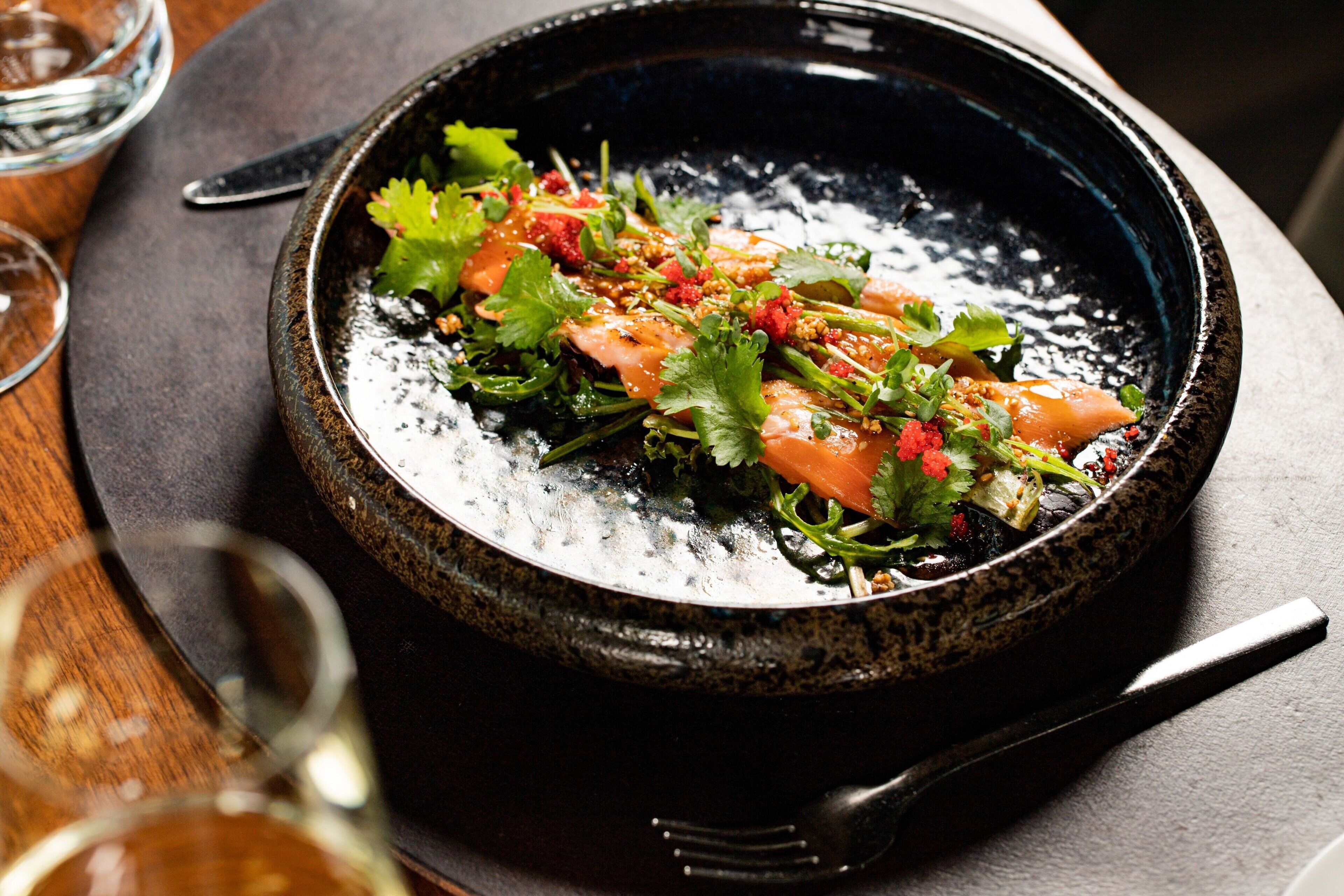 Un bol en céramique noire contenant un plat de saumon, garni d’herbes et d’épices, posé sur une surface sombre, avec une fourchette à côté.