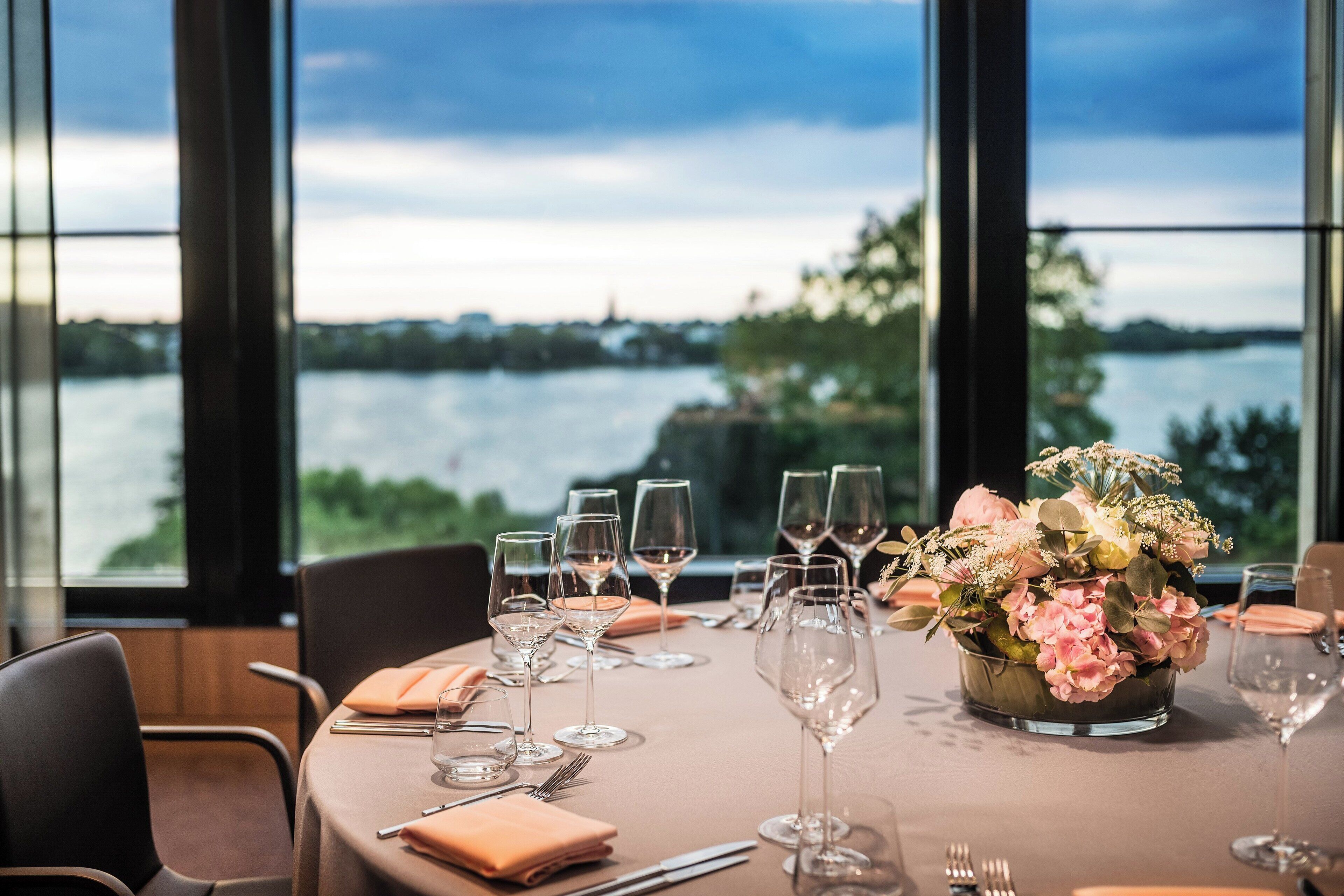 A table set for a meal with wine glasses and a floral centerpiece, overlooking a body of water.